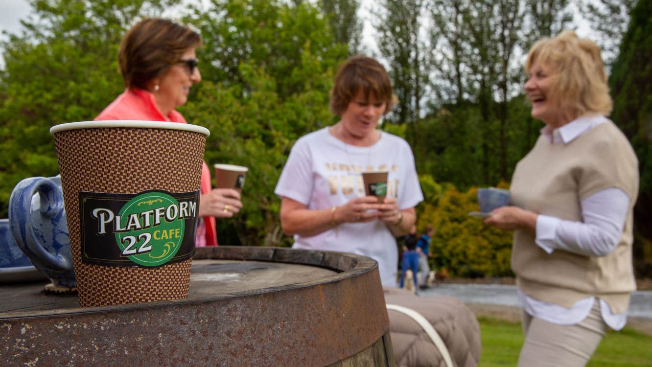 Three ladies outside having coffee with a take away coffee cup on a barrel to the forefront