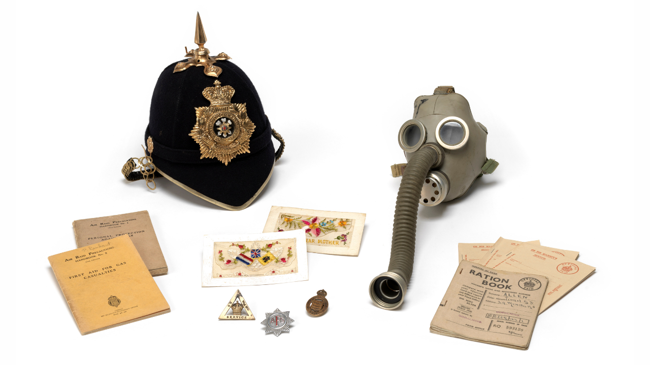 Against plain white background, various items from museum are laid out including a black helmet with gold brooch and point on the top, an grey gas mask, old ration books, small brooches and cards.