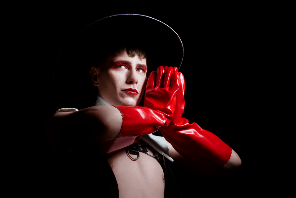 A person with a black rimmed hat, bare chested has raised arms with hands palms together in front of them, wearing red, shiny, plastic gloves. They are looking to their right, all against a dark background.