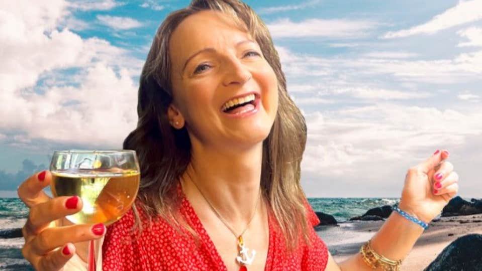 A laughing woman holding a glass of white wine with beach in the background.