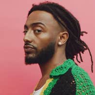 A man is looking sideways at the camera in green crocheted top against dull pink background.