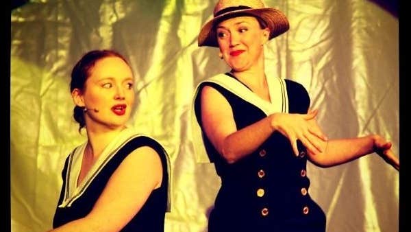 2 people in dark sailor style dresses are performing with gold background.