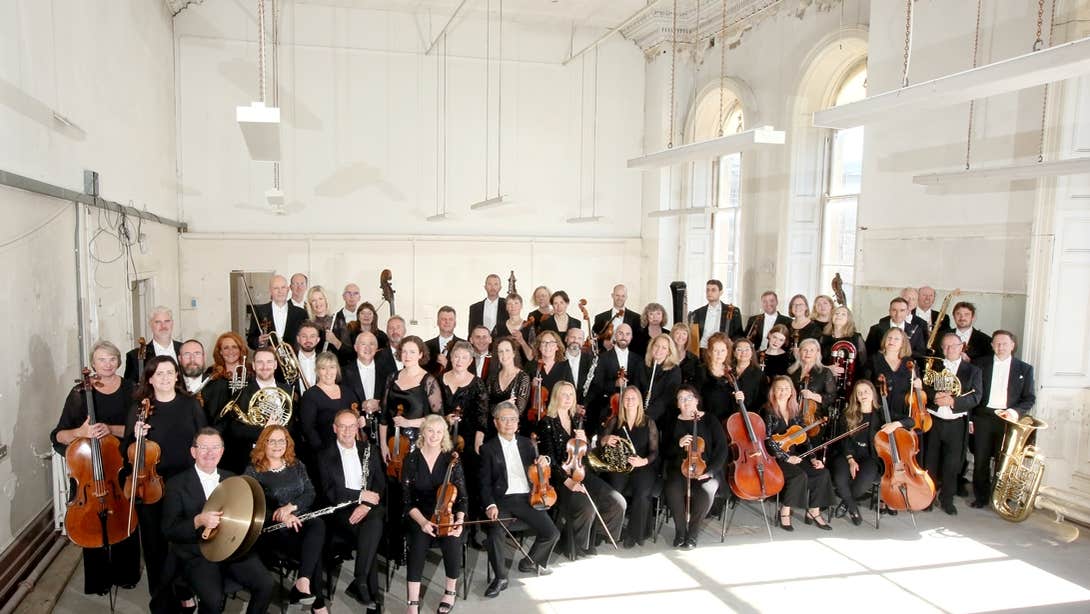 National Symphony Orchestra Ireland, a large orchestra posing in a group with their instruments in a large white room.