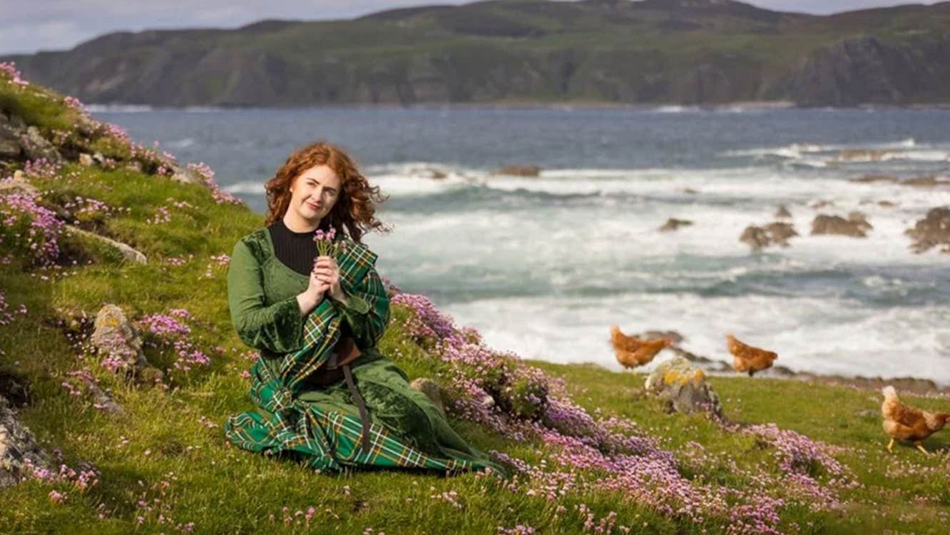 Person dressed in green tartan sitting among pink wildflowers on a grassy hillside near the sea