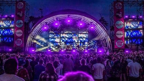 A large, outside, festival stage viewed from back in the audience, lit with purple lights at night in the dark.