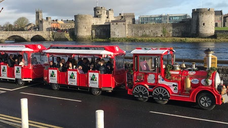 A small train on the road with a castle and the sea in the background