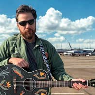 A bearded man in sunglasses is holding an ornately decorated black guitar with an airport and planes in the background.