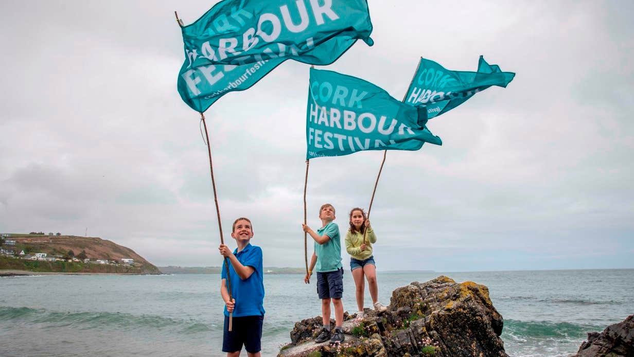 Photo of 3 children standing on large rocks by the edge of the sea, waving large teal coloured flags with event details on in white and blue text.