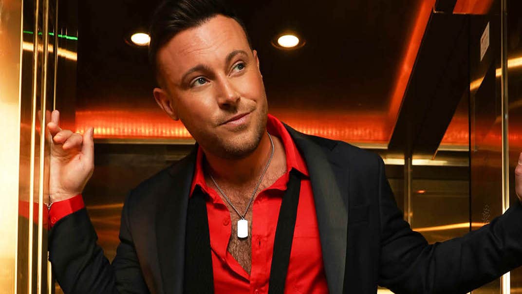 A man in a red open shirt and dark jacket appears to be looking out of the entrance of a small, gold lift, looking slightly bemused.