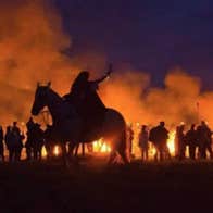 Person on a horse and line of figures at night silhouetted against flames and red smoke.