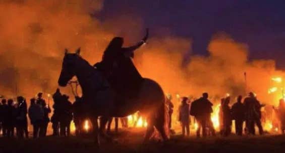 Person on a horse and line of figures at night silhouetted against flames and red smoke.