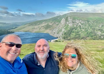 A couple and a tour guide posing for a selfie with a scenic background of a lake and mountains