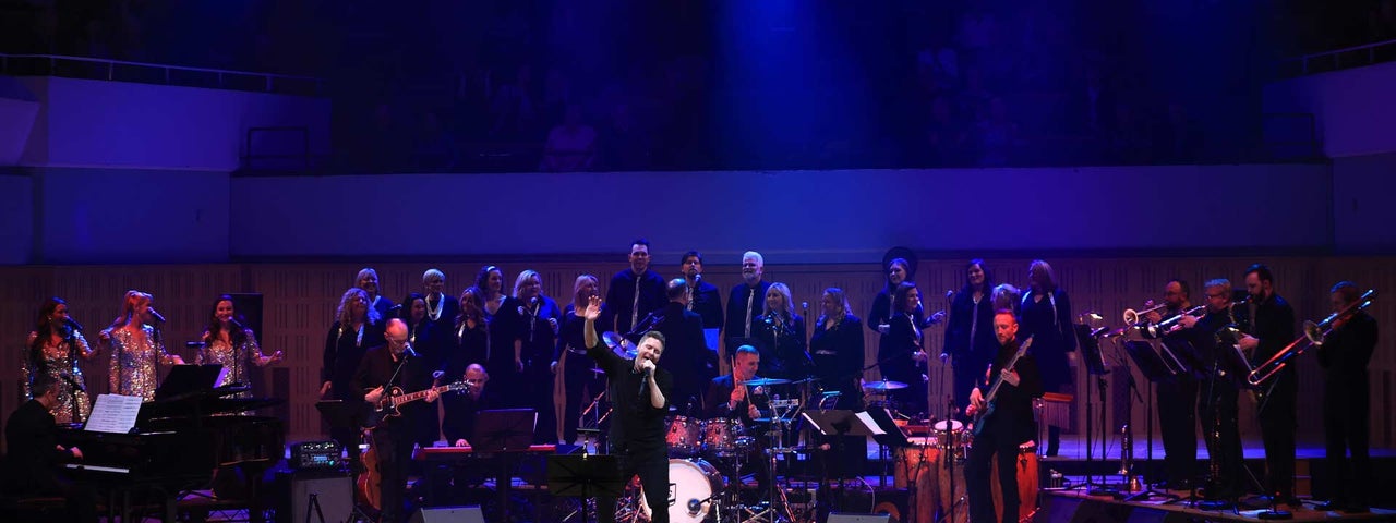 A man singing on a large stage with large band and choir behind him.