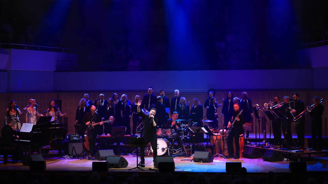 A man singing on a large stage with large band and choir behind him.