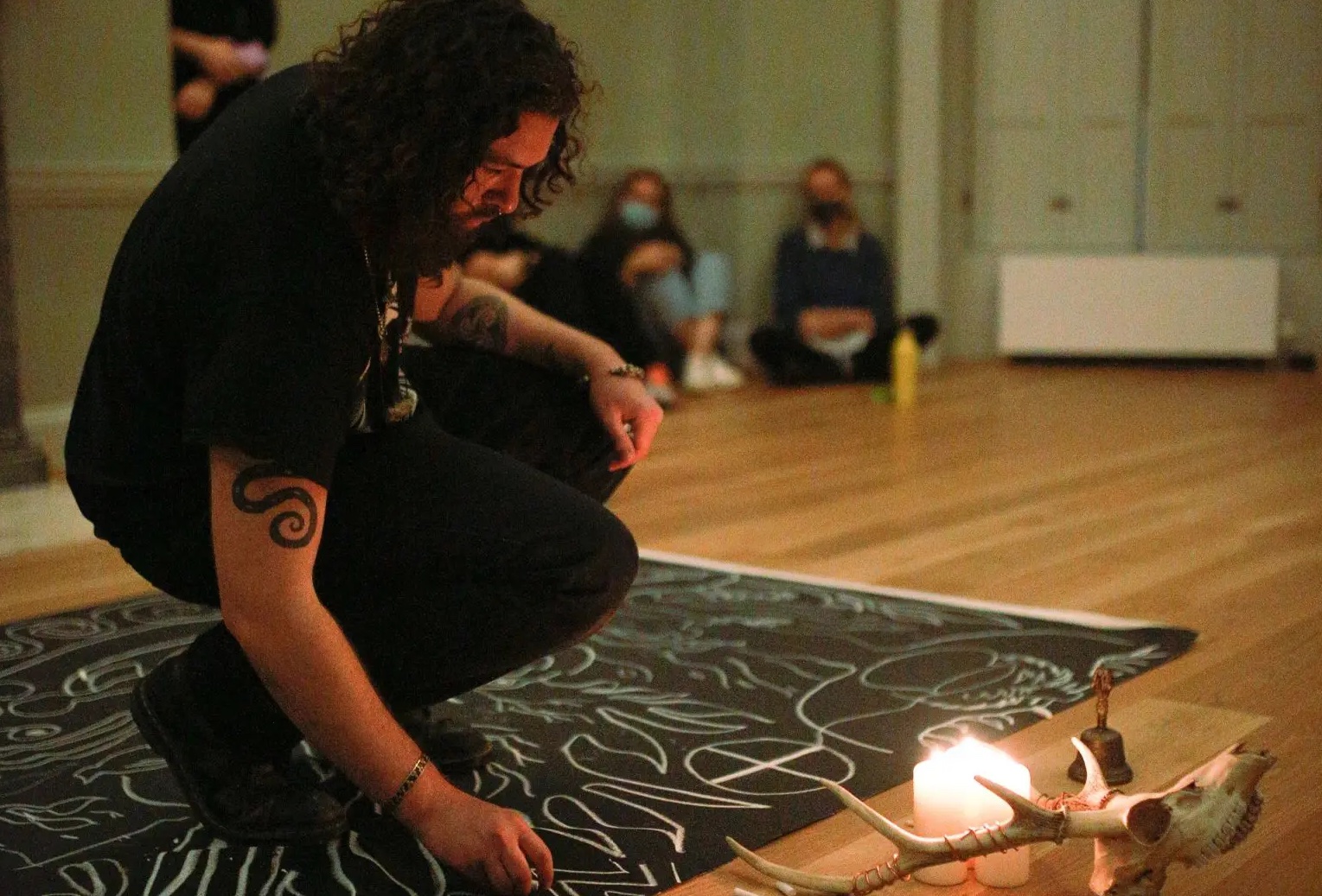 A man dressed in dark clothes in crouching down on a black mat with white designs on it with lit candles and a set of antlers & skull beside, all in a room on a wooden floor with a couple of people in the background.