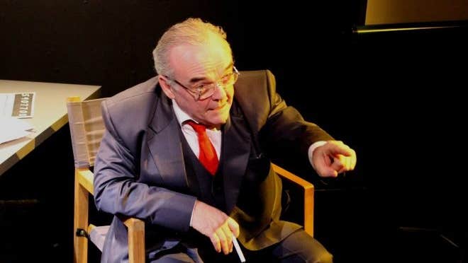 An older man in a suit is seated in a chair, talking and pointing with left finger