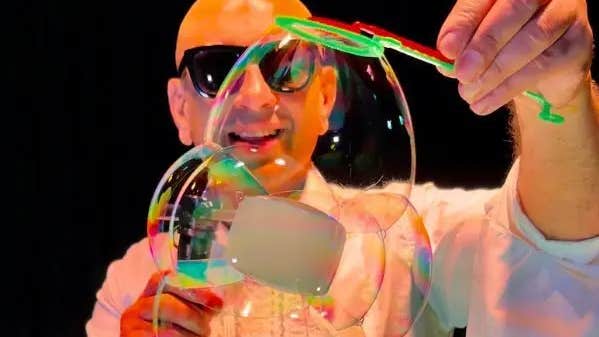 A smiling, bald man wearing sunglasses is holding an interlinked display of large bubbles, lit with orange light against black background.