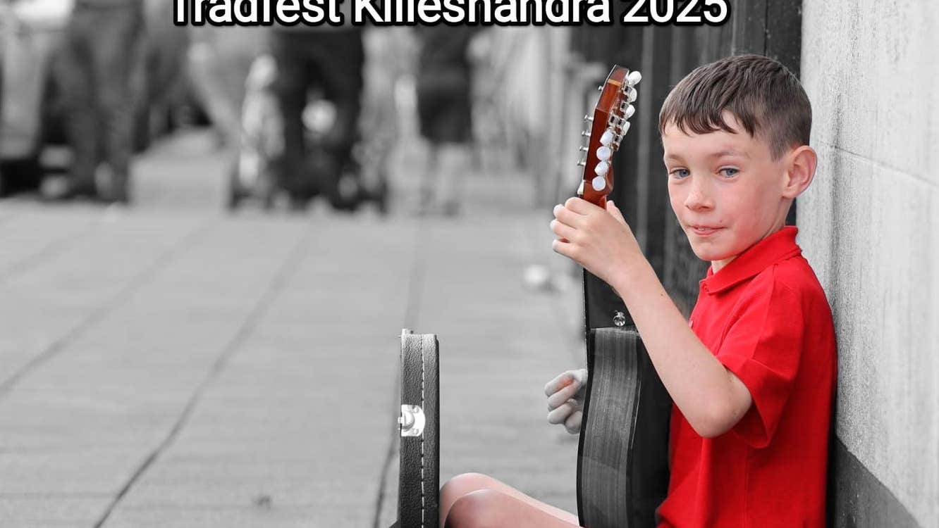 A young lad in bright red top is seated on a grey pavement leaning against a grey wall with a stringed instrument he is playing.