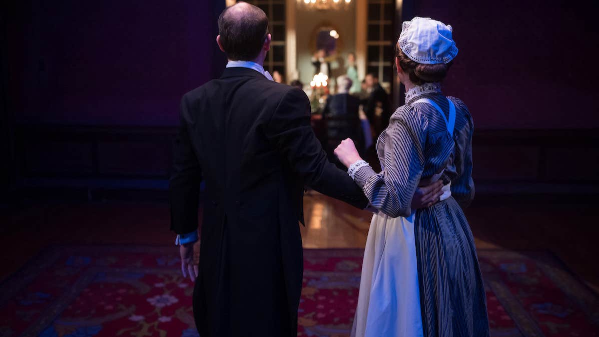 Two characters look into a brightly lit dining room where other characters are.