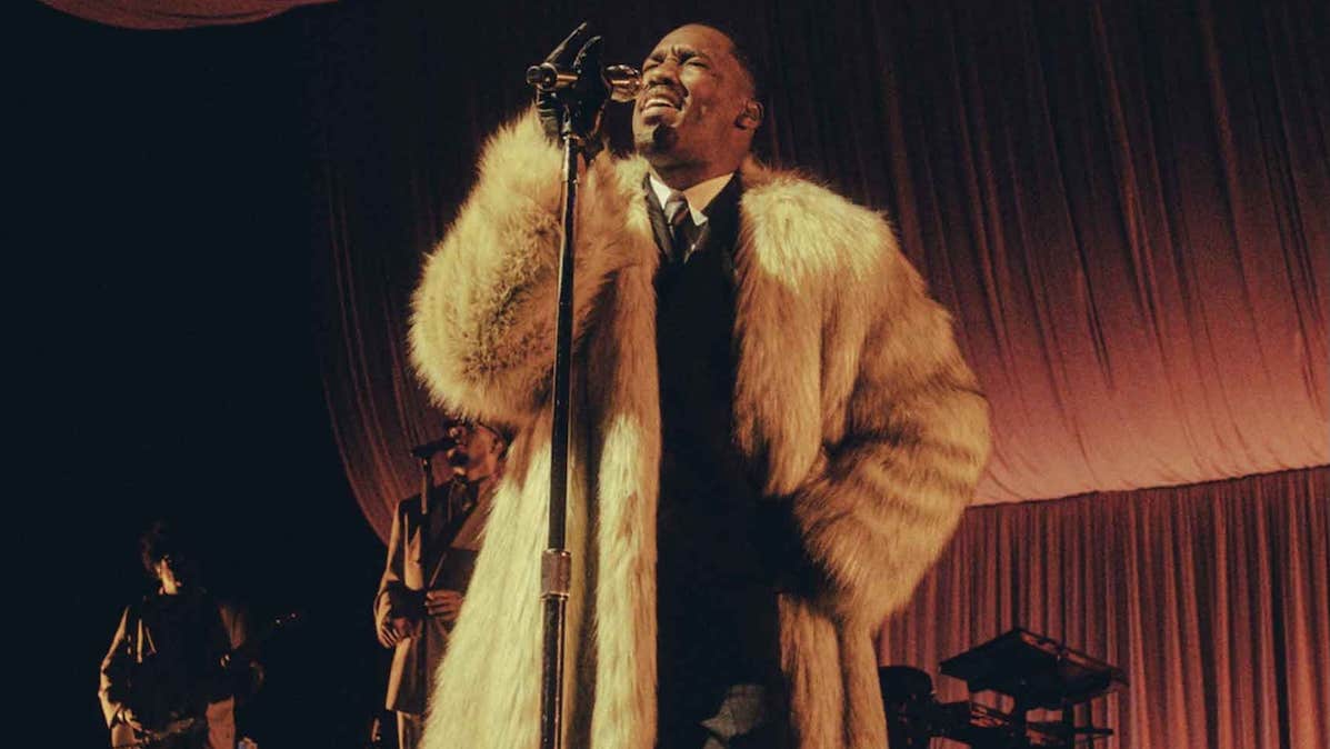 R&B superstar GIVĒON, a man in large, light coloured fur coat singing into a mic on a stand.