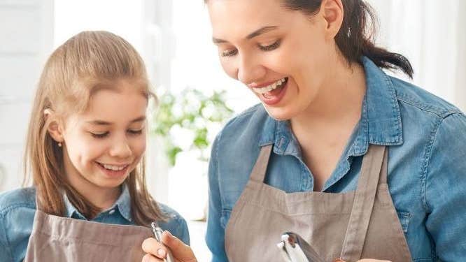 A woman and young girl wearing identical aprons are smiling, looking down at steaming saucepan on a stove