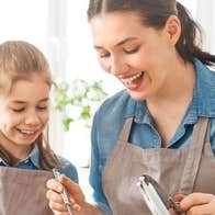 A woman and young girl wearing identical aprons are smiling, looking down at steaming saucepan on a stove