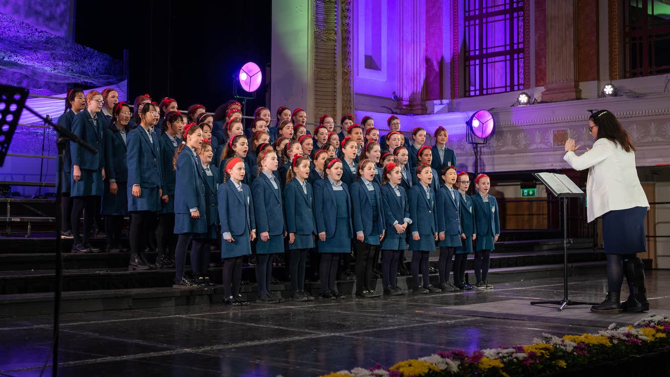 Children in school uniform standing on a stage in tiered rows singing with conductor in front.