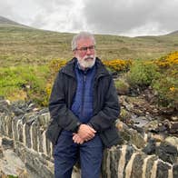 An elderly Gerry Adams, sitting on a wall in the wilderness.