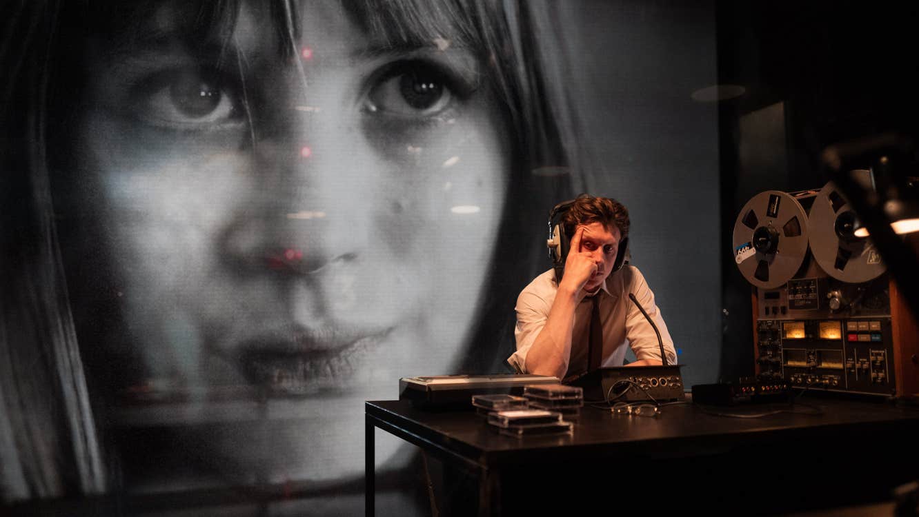 A man at a desk with audio equipment. An image of Marianne Faithful is being projected behind him.