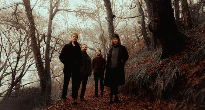 4 men in dark clothing standing in a wooded area in winter.