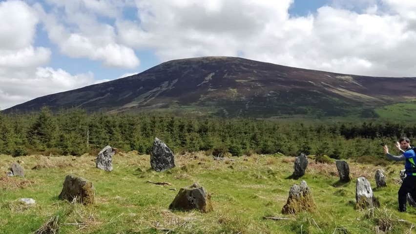 Man standing in front of standing stone circle with hands outstretched set against a wooded and mountain backdrop