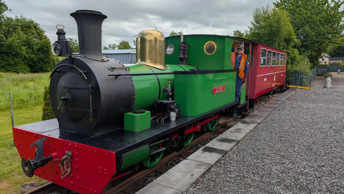 A small green steam engine at a platform pulling a single red passenger carriage
