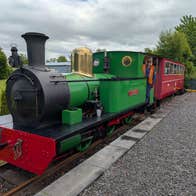 A small green steam engine at a platform pulling a single red passenger carriage