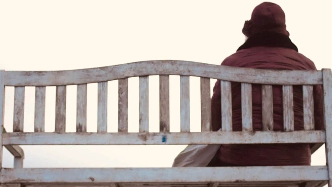 'An tÉalú' Ceoldráma Nua le Seán Óg Ó Duinnín - rear view of person in winter coat and hat seated at one end of a wooden bench.