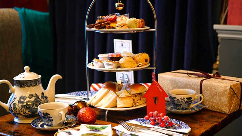Festive Afternoon Tea stand displayed on a table in the Old Music Shop Restaurant, featuring scones, sandwiches, brioche buns, mince pies, miniature desserts, cups of tea, and a gift box, surrounded by Christmas decorations and ornaments.