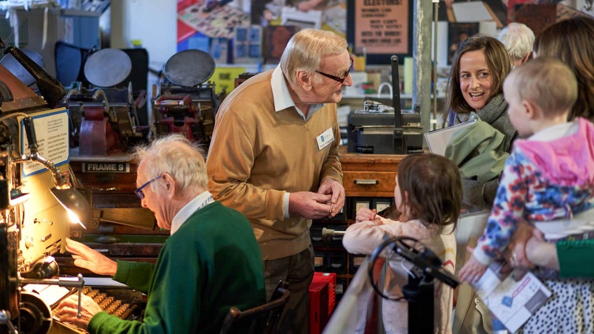 2 older men talking and demonstration old equipment to a small group of adults and children.