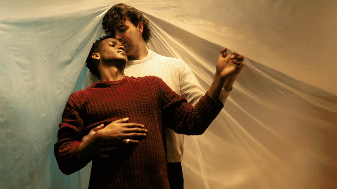 2 people in a loving embrace, one standing behind the other, against a cloth background.