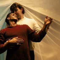 2 people in a loving embrace, one standing behind the other, against a cloth background.