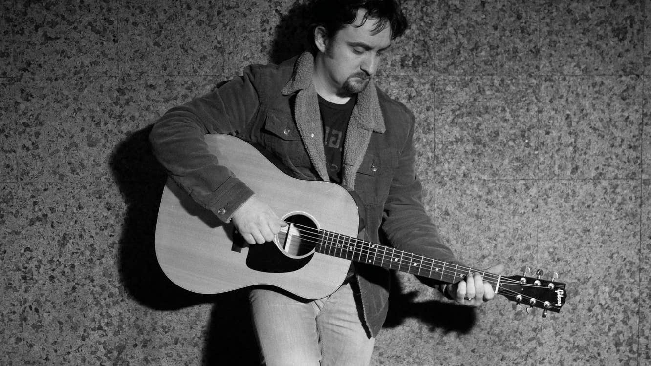 A man leaning against a wall playing a guitar looking down