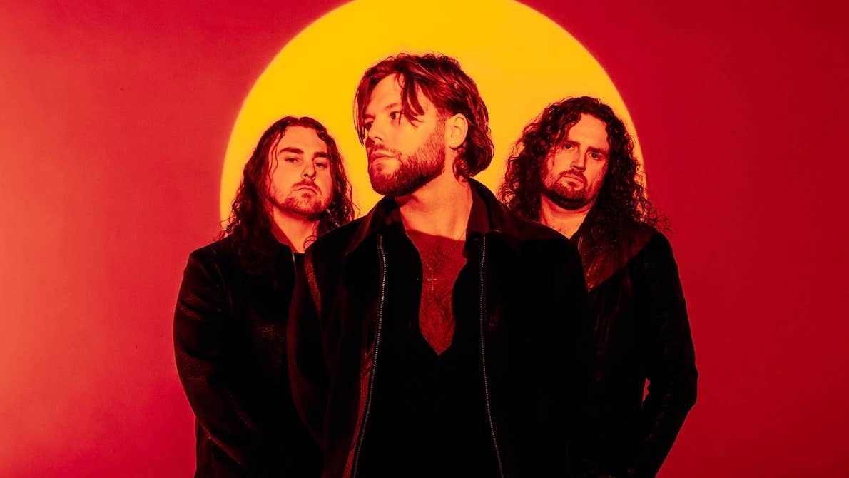 Rock band Florence Black, 3 moody looking men dressed in black with orange full sun background against red