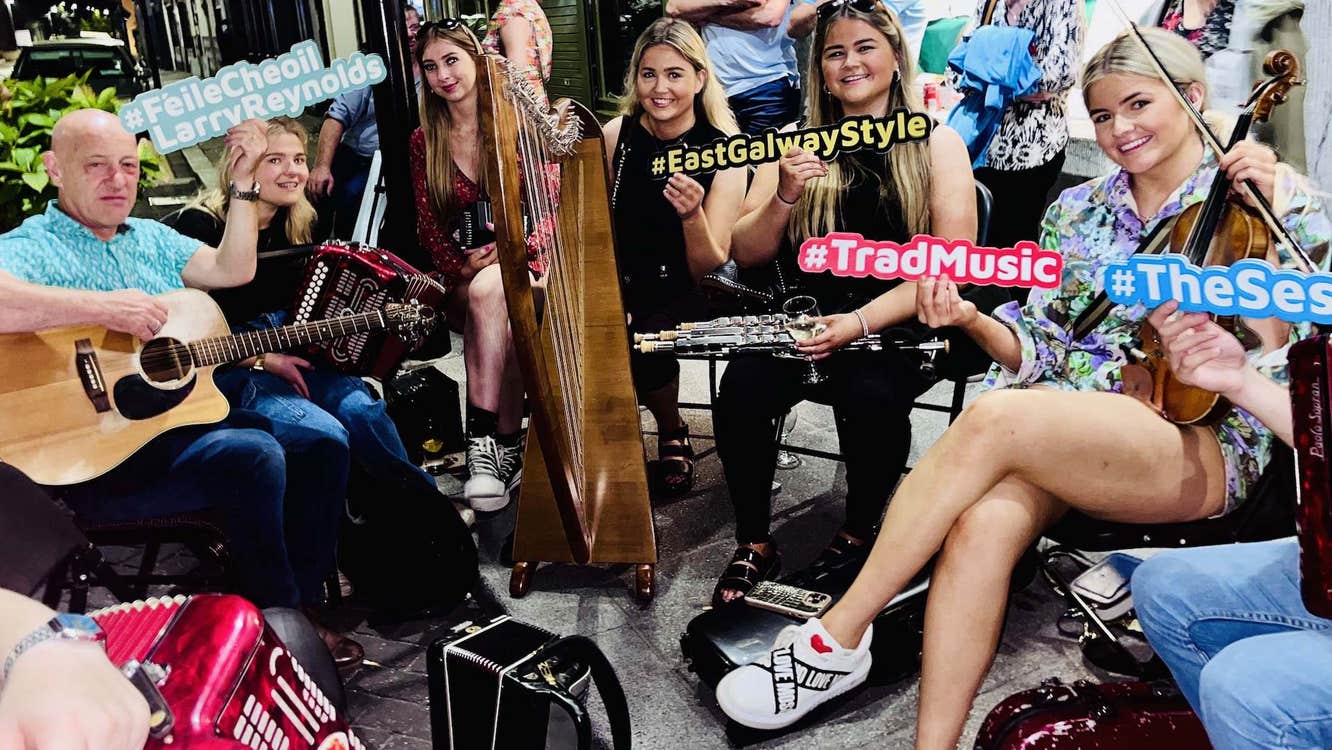 A group of musicians with instruments are seated in a close group on a pavement, some holding different coloured hash tag signs, smiling at the camera.