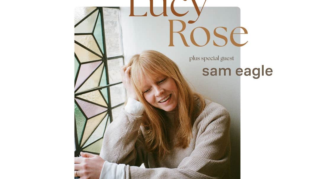 British songwriter Lucy Rose, a smiling woman leaning her head on one hand, looking downwards.