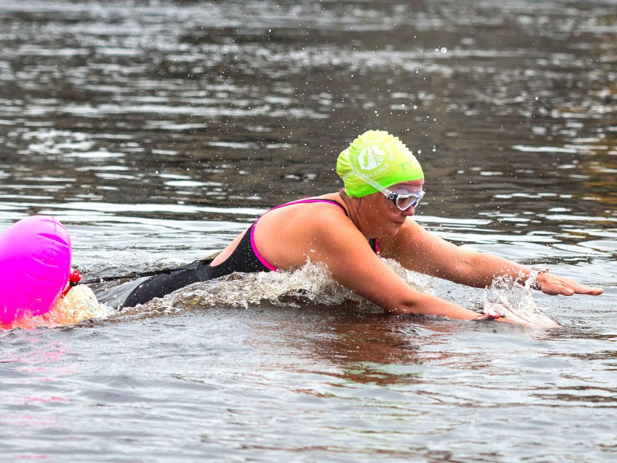 A woman in water swimming with a bright green swim hat and goggles on, with the pink covered head of another swimmer beside her.