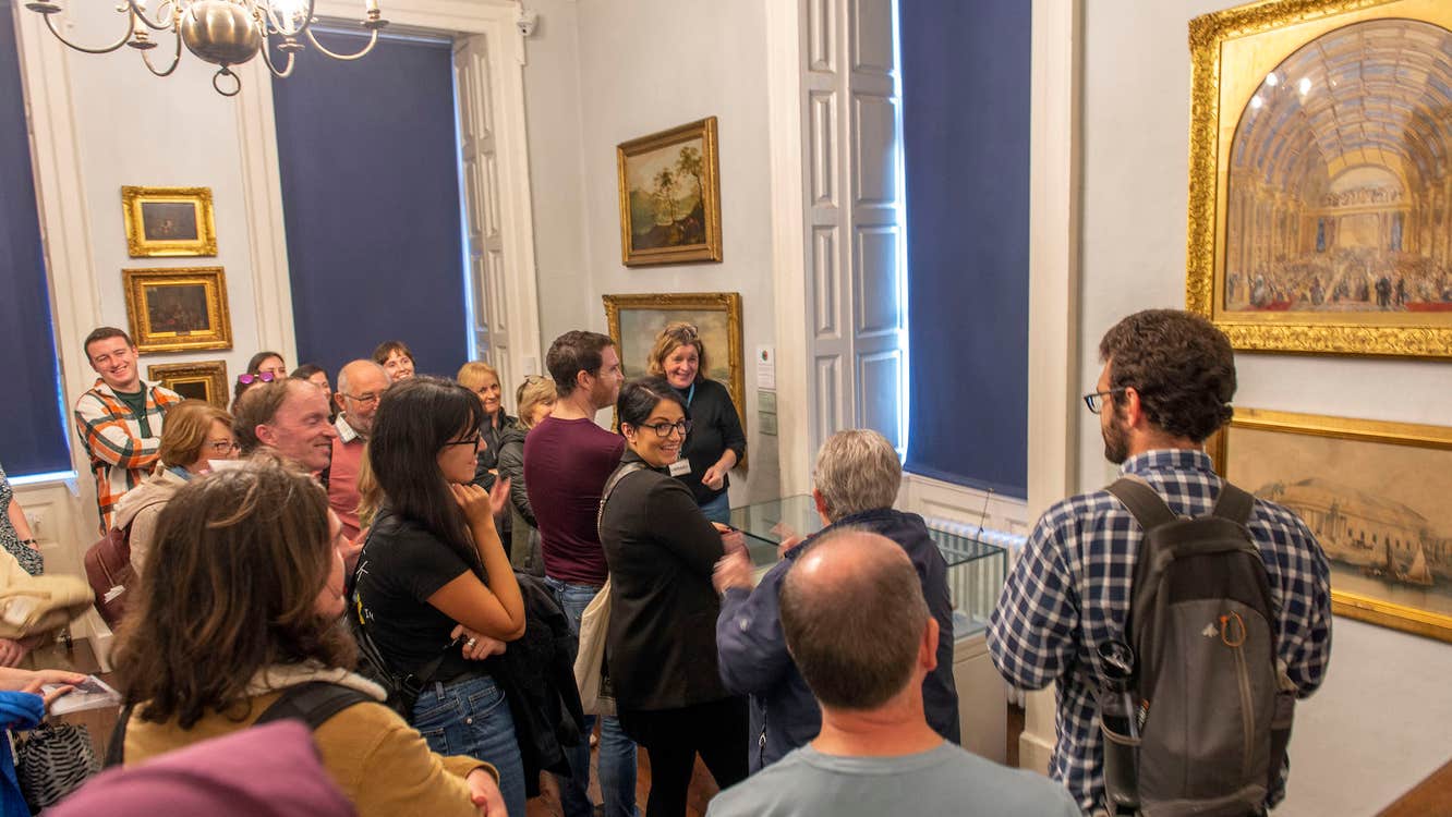 A group of people in a gallery are gathered round a small glass cabinet, listening to a tour guide speak.