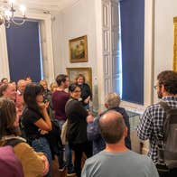 A group of people in a gallery are gathered round a small glass cabinet, listening to a tour guide speak.
