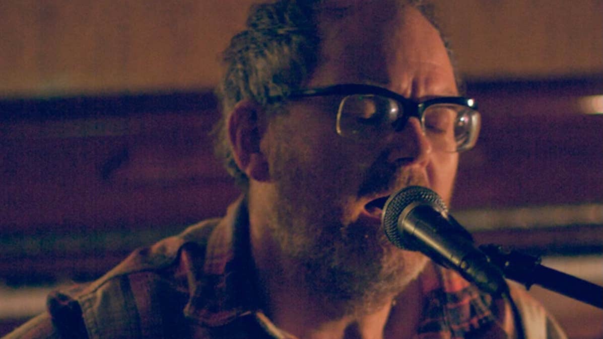 A darkly lit close up of man singing into a mic on a stand.