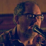 A darkly lit close up of man singing into a mic on a stand.