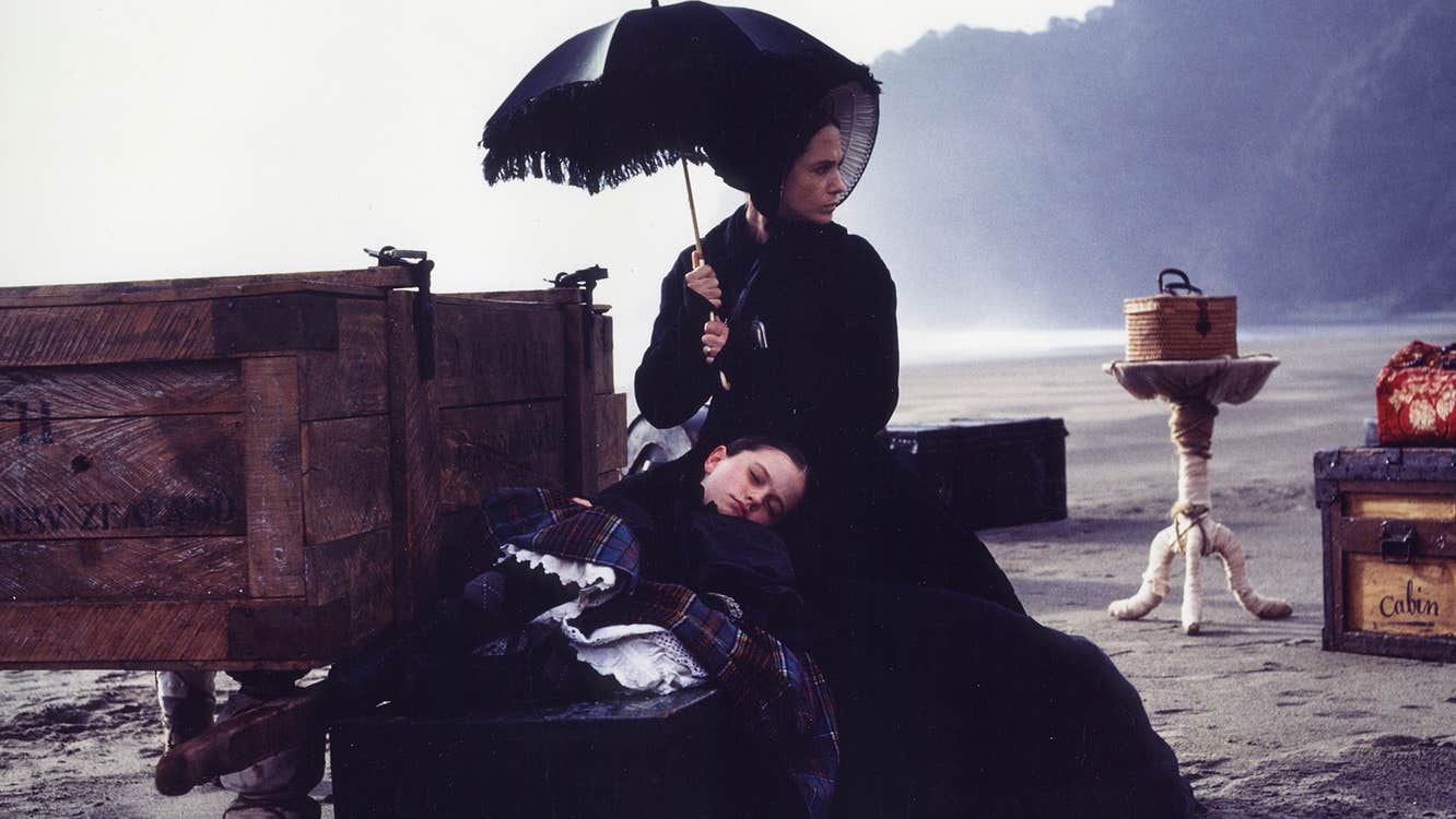 A girl resting against the lap of a woman with an umbrella. They are surrounded by luggage on a beach.