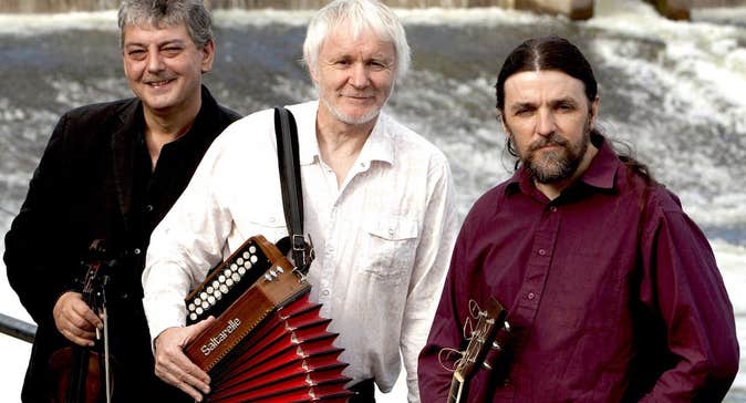 Music Network presents Máirtín O’ Connor Trio, 3 men holding musical instruments standing against railings with cascading water behind them.