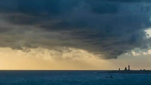 View the horizon over a flat, dark sea with dark clouds and a lighthouse in the distance.